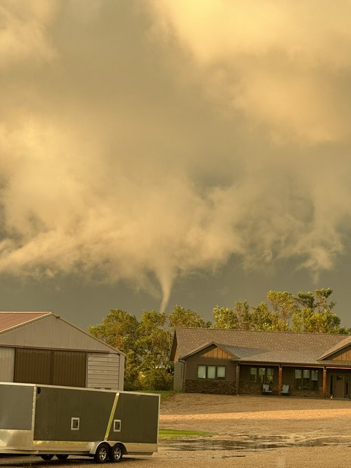 Home, garage, and trailer in the foreground. A tornado is in the center of the image, descending from a white cloud base into trees behind the house.